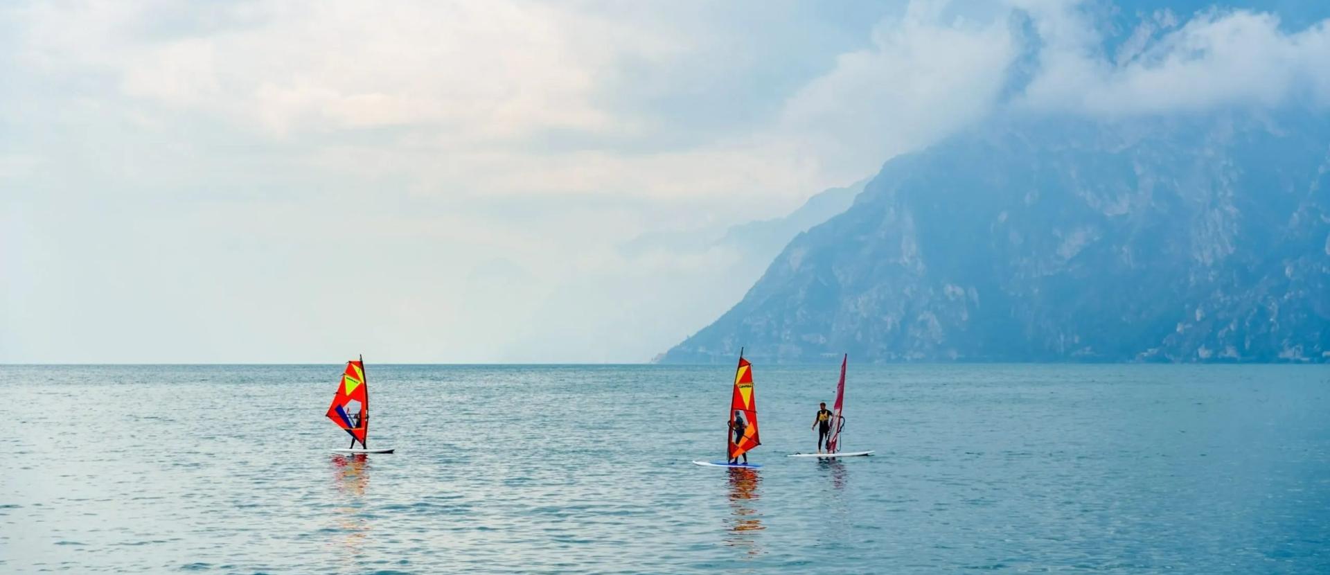 Windsurfisti solitari solcano acque calme, montagne avvolte da nubi in lontananza.
