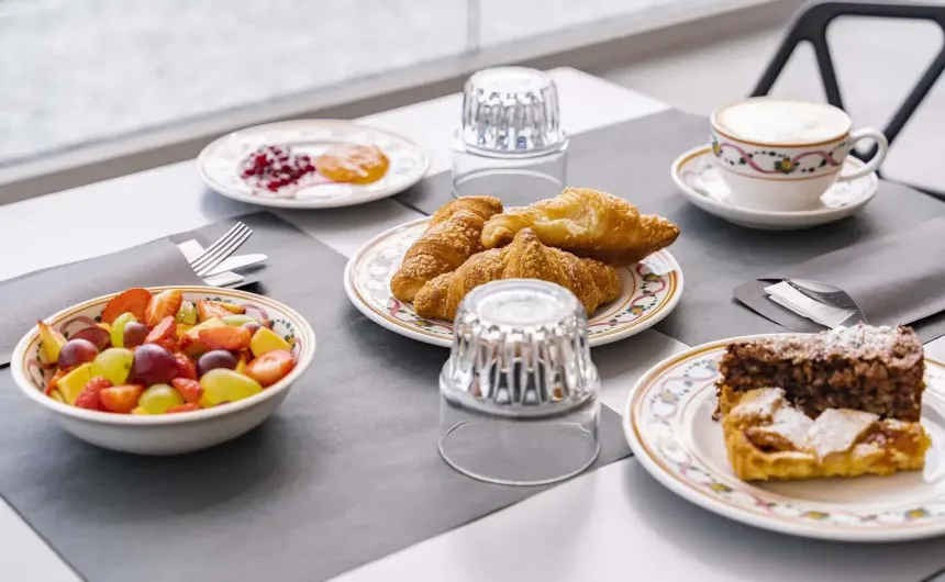 Colazione elegante: croissant, macedonia, torte, marmellate e cappuccino su tavola raffinata.