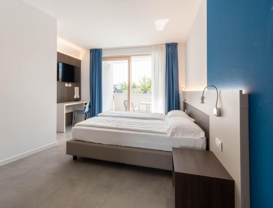Camera luminosa, moderna, con letto matrimoniale, tende blu e balcone con vista verde.