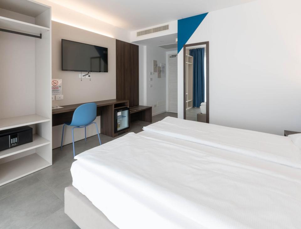 Camera moderna, luminosa e minimalista con letto bianco, scrivania e TV a parete.