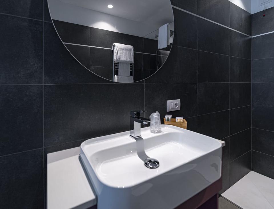 Elegante bagno moderno con pareti scure, lavabo bianco e grande specchio rotondo.