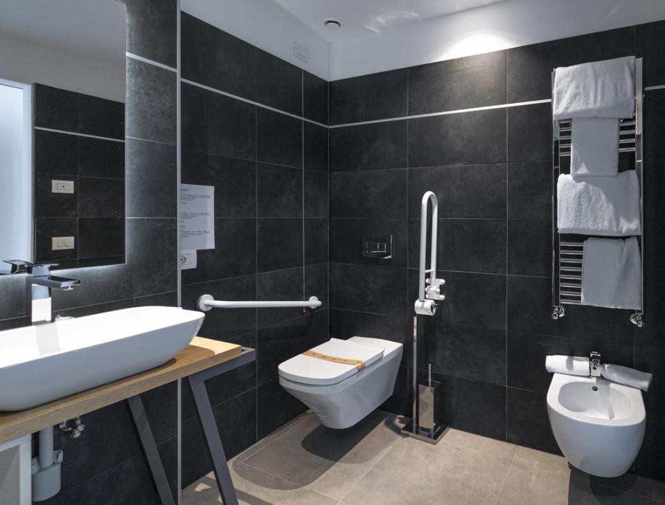 Bagno moderno, accessibile, con piastrelle scure, lavandino elegante e asciugamani bianchi.
